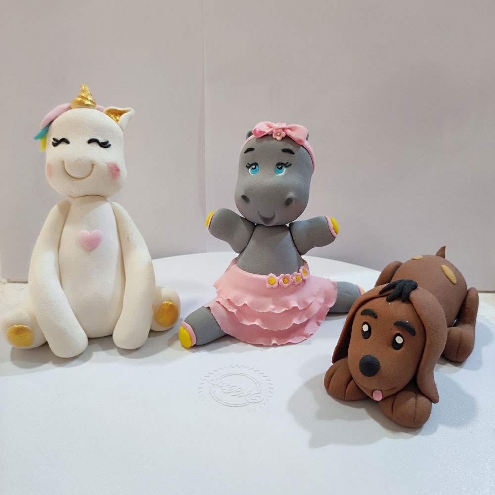 Figuras de fondant modeladas: un unicornio blanco con cuerno dorado, una hipopótamo bailarina con vestido rosa, y un perro marrón con orejas largas y expresivas